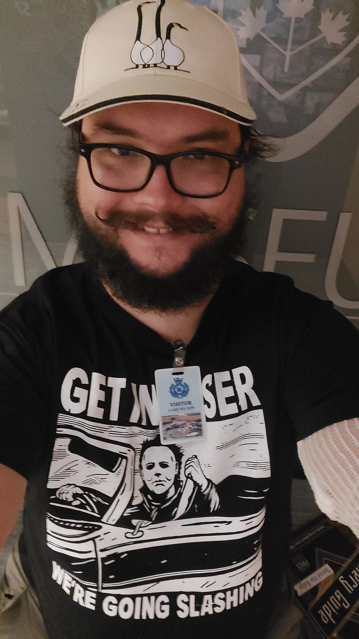 A man with short hair and bushy facial hair stands in front of a large Ontario Provincial Police emblem inside the OPP Museum wearing a stylaized Halloween (1978) t-shirt that reads "Get in Loser, We're Going Slashing", he is also wearing a clip on visitor's badge for the museum